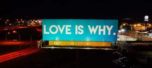 Blue billboard saying Love is why placed on road surrounded by cars and street lights against black night