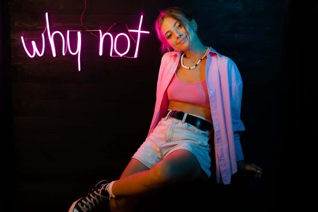 Young woman posing indoors under pink neon 'why not' sign, 
 freelancer expressing individuality.